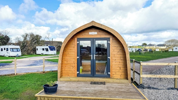 A small wooden structure with a glass door, surrounded by a gravel path and grass. - Lavender is a cosy pod on the site of the Centry Touring Park in Brixham. (Brixham)