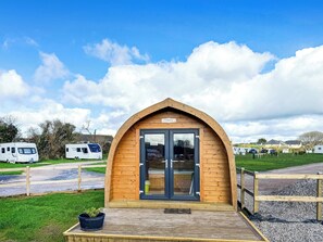 A small wooden structure with a glass door, surrounded by a gravel path and grass. - Lavender is a cosy pod on the site of the Centry Touring Park in Brixham. (Brixham)