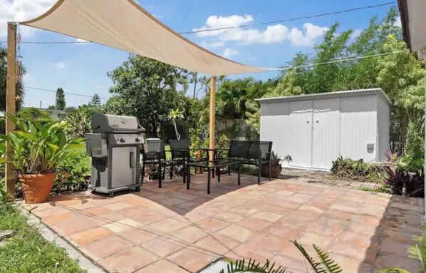 Enjoy al fresco dining under a spacious canopy, surrounded by lush greenery and a barbecue grill.