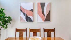 Une table à manger avec trois chaises en bois et un saladier rempli de fruits. Deux tableaux abstraits sont accrochés au mur, au-dessus de la table.