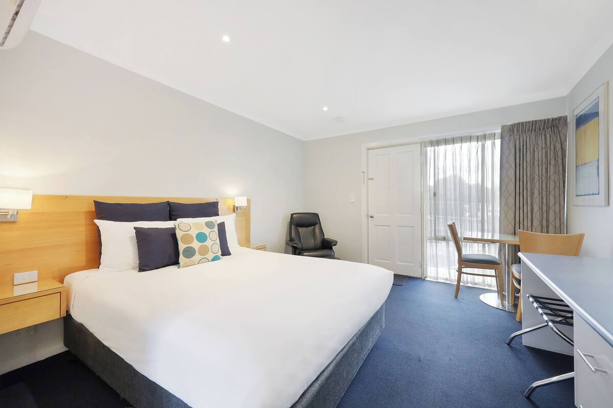 A hotel room with a large bed, a desk, a chair, and a door leading to a balcony.