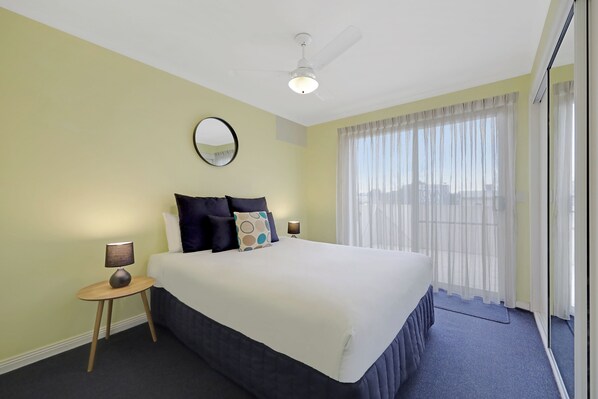 A hotel room with a large bed, a round mirror, a ceiling fan, and a view through sheer curtains.