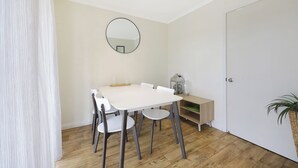 Uma sala de jantar com uma mesa e cadeiras brancas, um espelho redondo na parede, piso de madeira e uma porta branca.