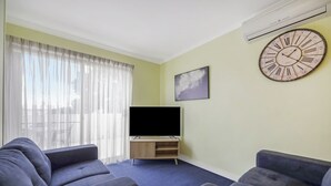 Uma sala de estar com dois sofás, uma TV de tela plana sobre um suporte de madeira, um relógio de parede grande e uma janela com cortinas leves.