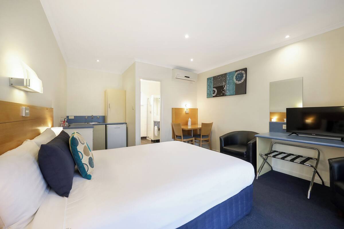 A hotel room with a large bed, a desk with a chair, a television, and a small kitchenette.