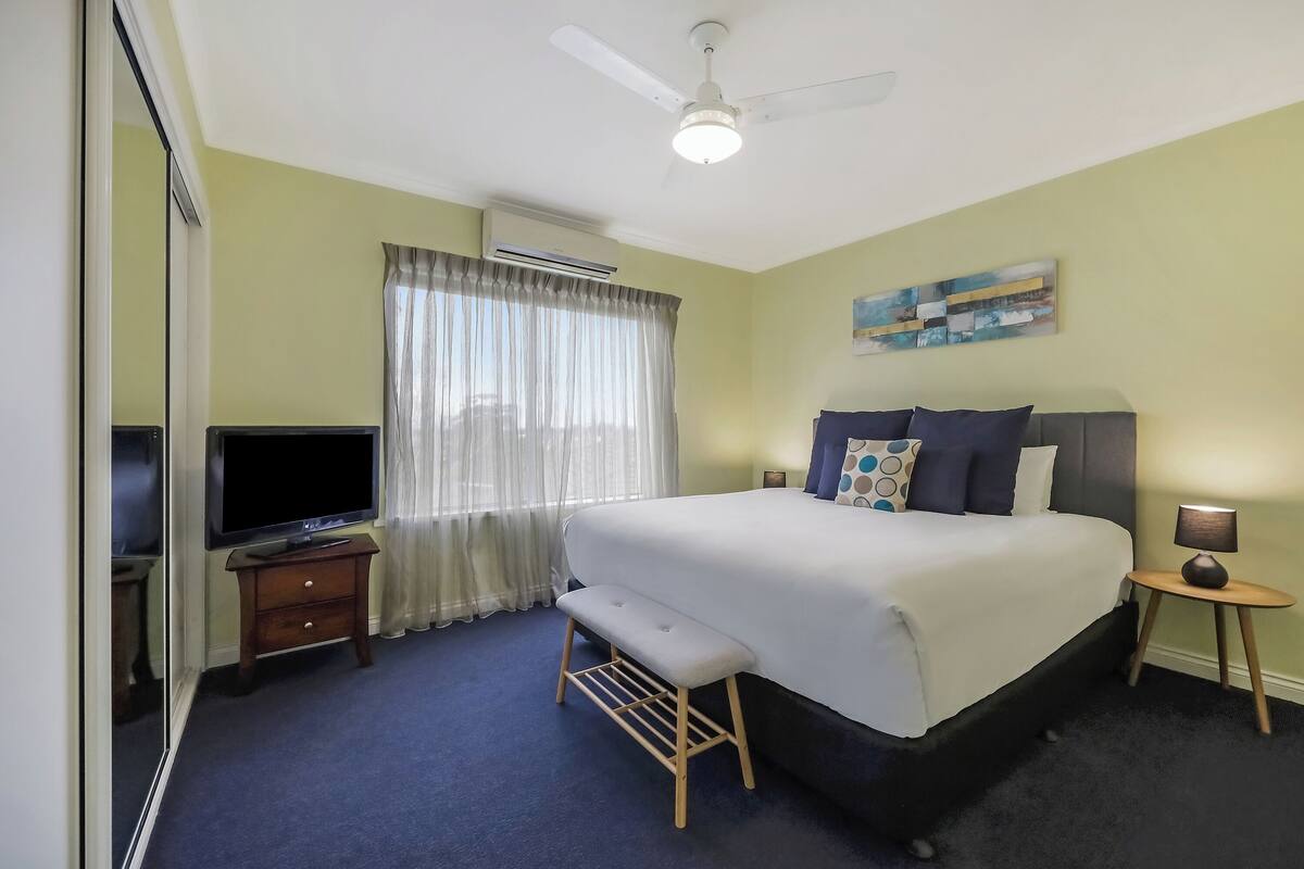 A hotel room with a bed, a TV, a bench, a small table with a lamp, and a ceiling fan.