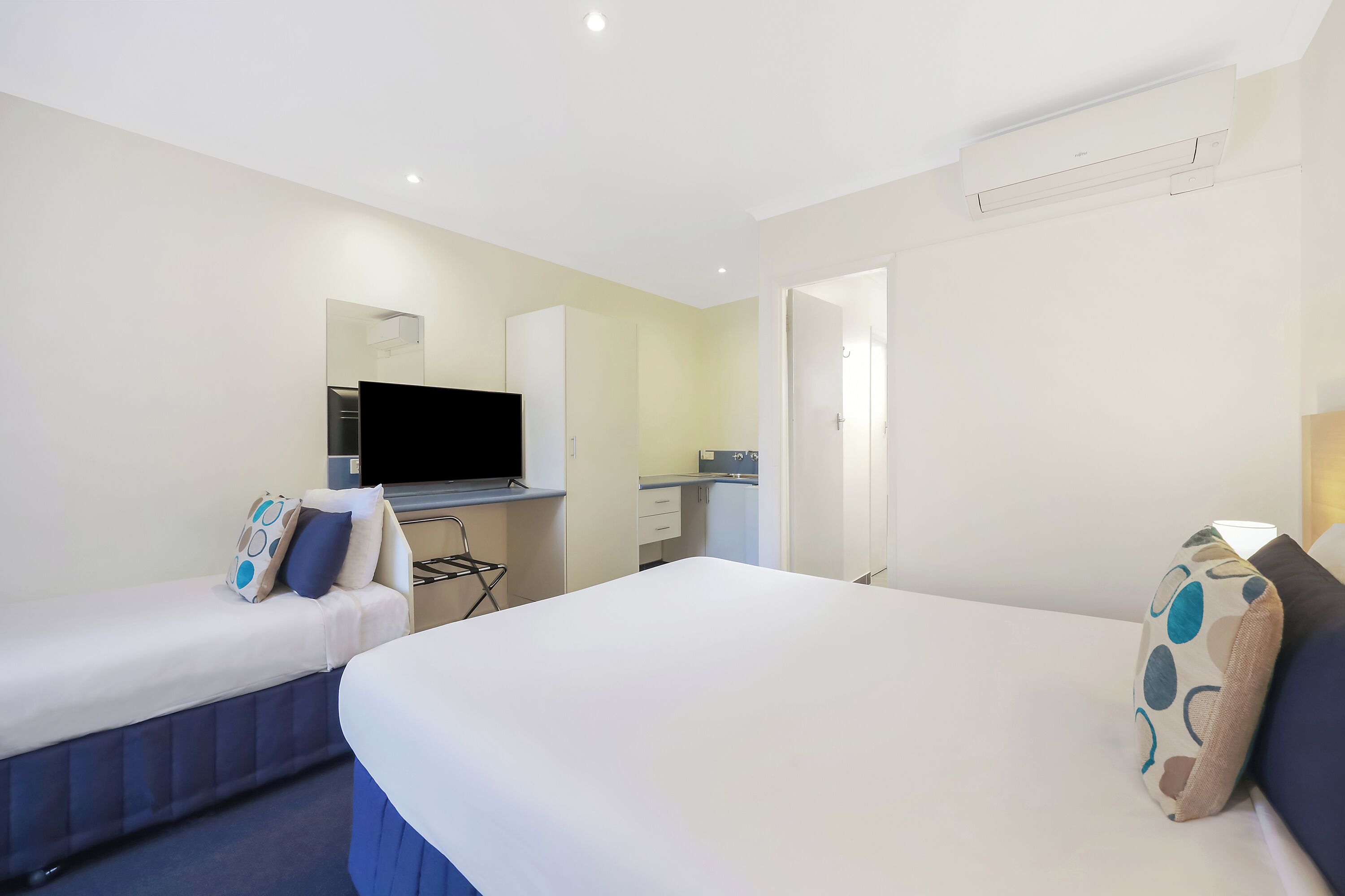 A hotel room with two beds, a TV, a desk, and a kitchenette.