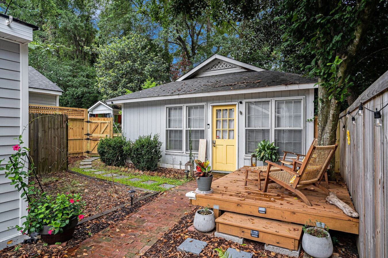 Una casita con terraza, una puerta amarilla y un jardín con plantas en macetas y una cerca de madera.