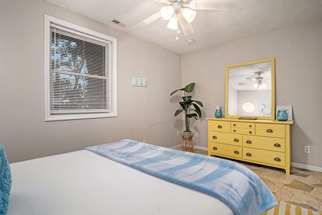 A bedroom with a bed, a dresser, a mirror, a ceiling fan, and a window with blinds.