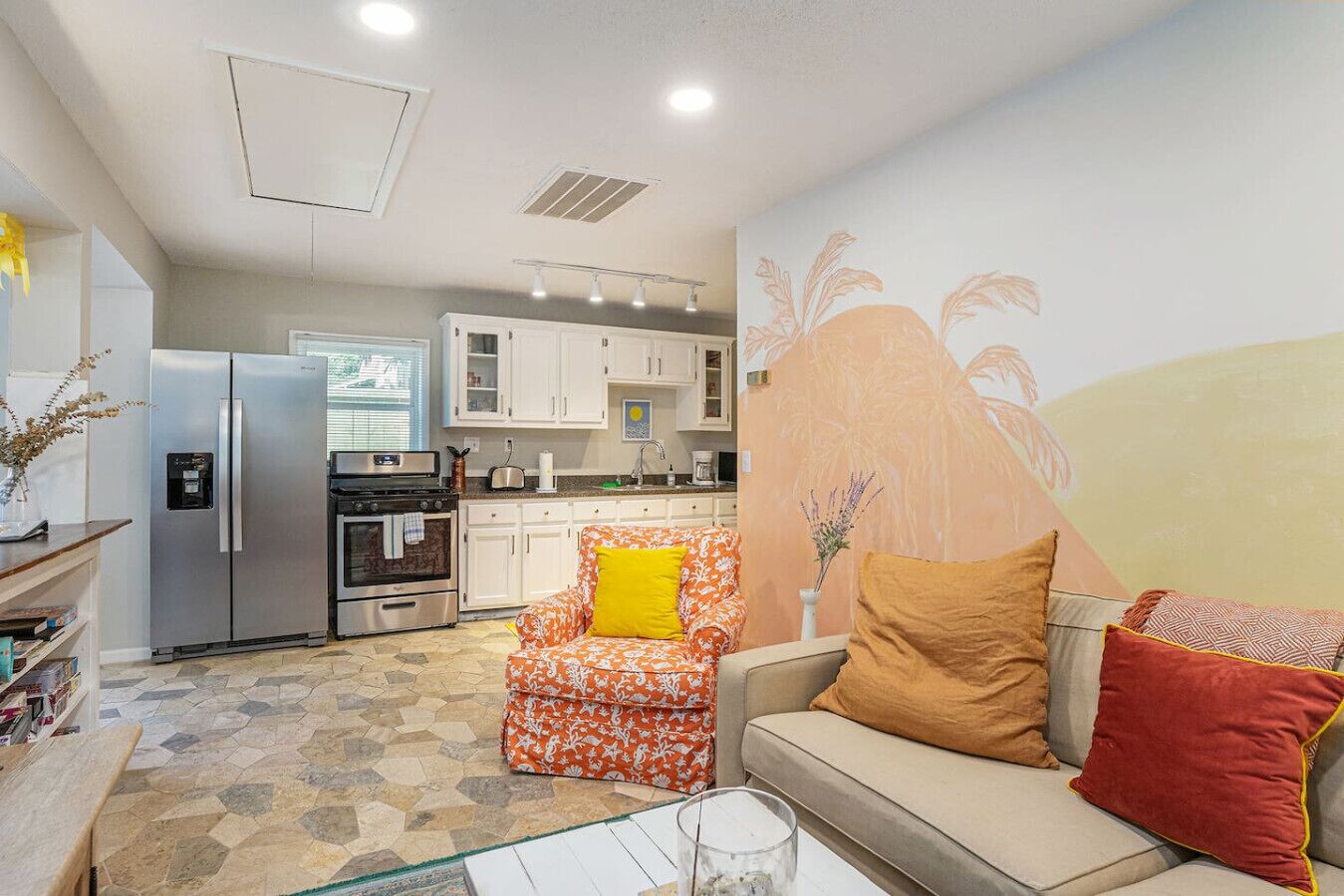 Espaço compacto com cozinha americana, sofá, poltrona estampada e um mural decorativo.