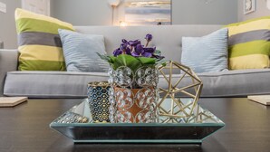 Un salon avec un canapé gris, des coussins décoratifs, une table basse en verre et un vase rempli de fleurs violettes.