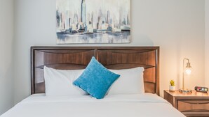 Un lit bien fait, avec du linge de lit blanc et un coussin bleu, une tête de lit en bois, un tableau représentant un paysage urbain accroché au mur, et une table de chevet avec une lampe et une petite plante.