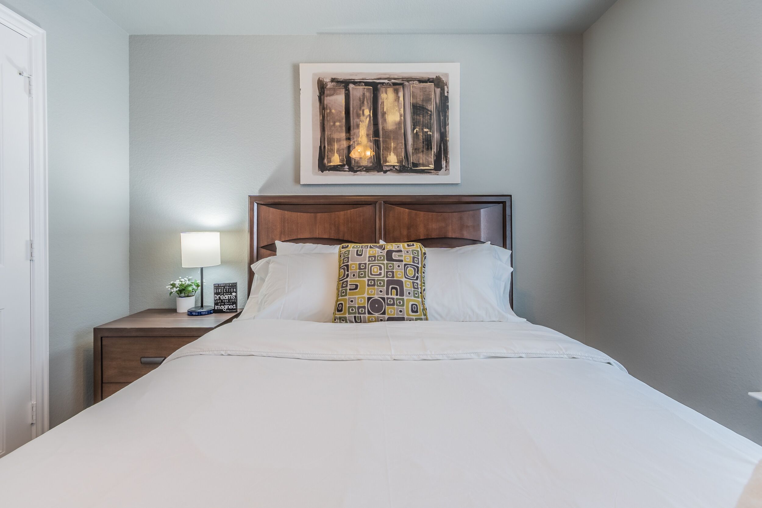 Una cama bien hecha con sábanas blancas, un cojín decorativo estampado y un cabecero de madera. Se ve una mesita de noche con una lámpara y una pequeña planta.