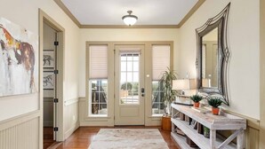 Un couloir avec un plancher en bois, des murs de couleur beige, une porte avec des stores, un miroir et une petite table ornée de plantes.