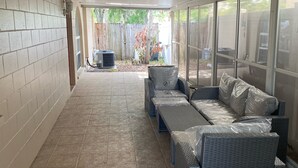 Un patio cubierto con suelo de baldosas, un conjunto de sofá de mimbre gris y una pared de ladrillo.