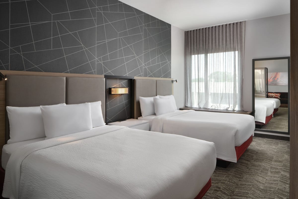 Ein Hotelzimmer mit zwei Betten, einem modernen Wanddesign im geometrischen Stil, einem großen Fenster mit transparenten Vorhängen und einem Spiegel an der Wand.