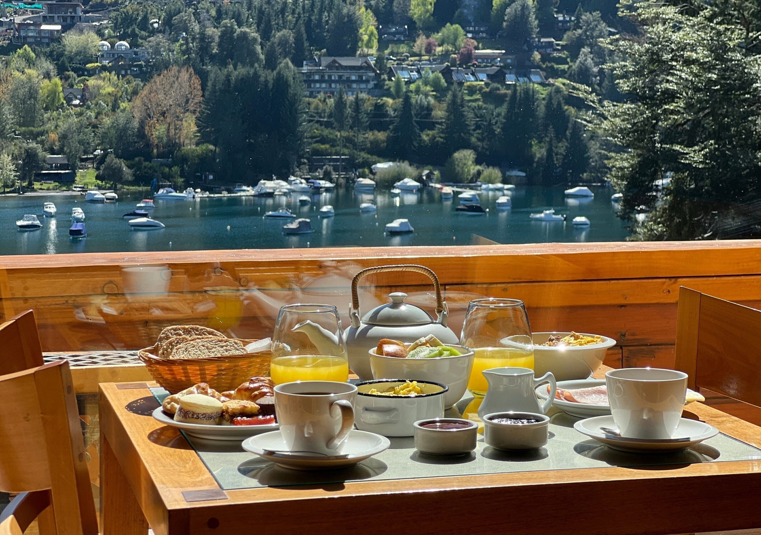 A breakfast tray with various food items and beverages on a table overlooking a lake with boats.