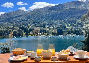 Una mesa de desayuno con diversas comidas y bebidas, con vistas a un lago con barcos y montañas al fondo.