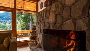 A stone fireplace with a fire, a wooden bench, and a view of the forest through a window. - El Muelle by DOT Boutique (Villa La Angostura)