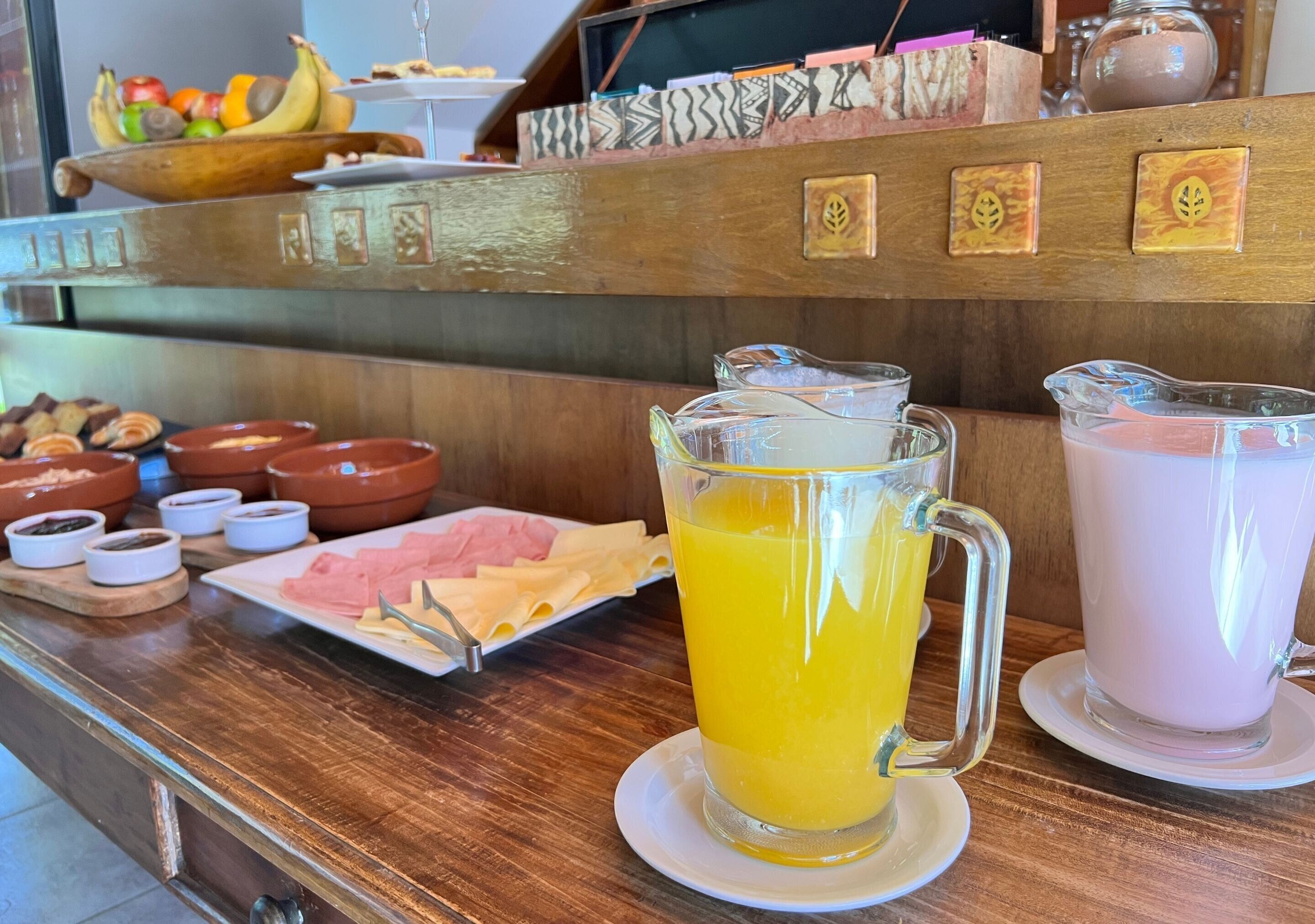 Una mesa de bufé con diversos alimentos, incluyendo una jarras de jugo de naranja y otra de una bebida blanca, posiblemente leche o jugo.