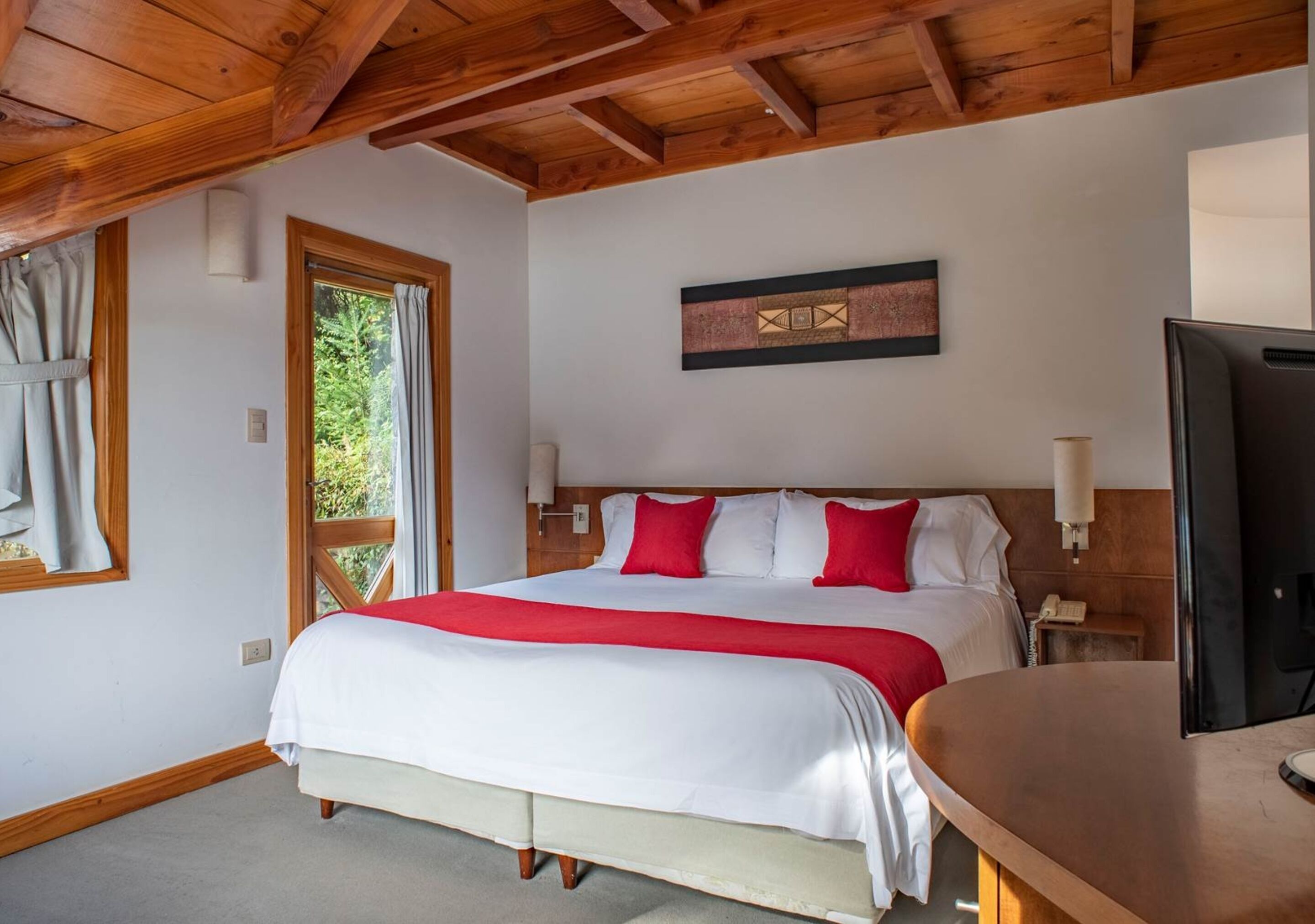 Une chambre à coucher avec un grand lit, des poutres apparentes en bois au plafond, un bureau et une fenêtre avec des rideaux.