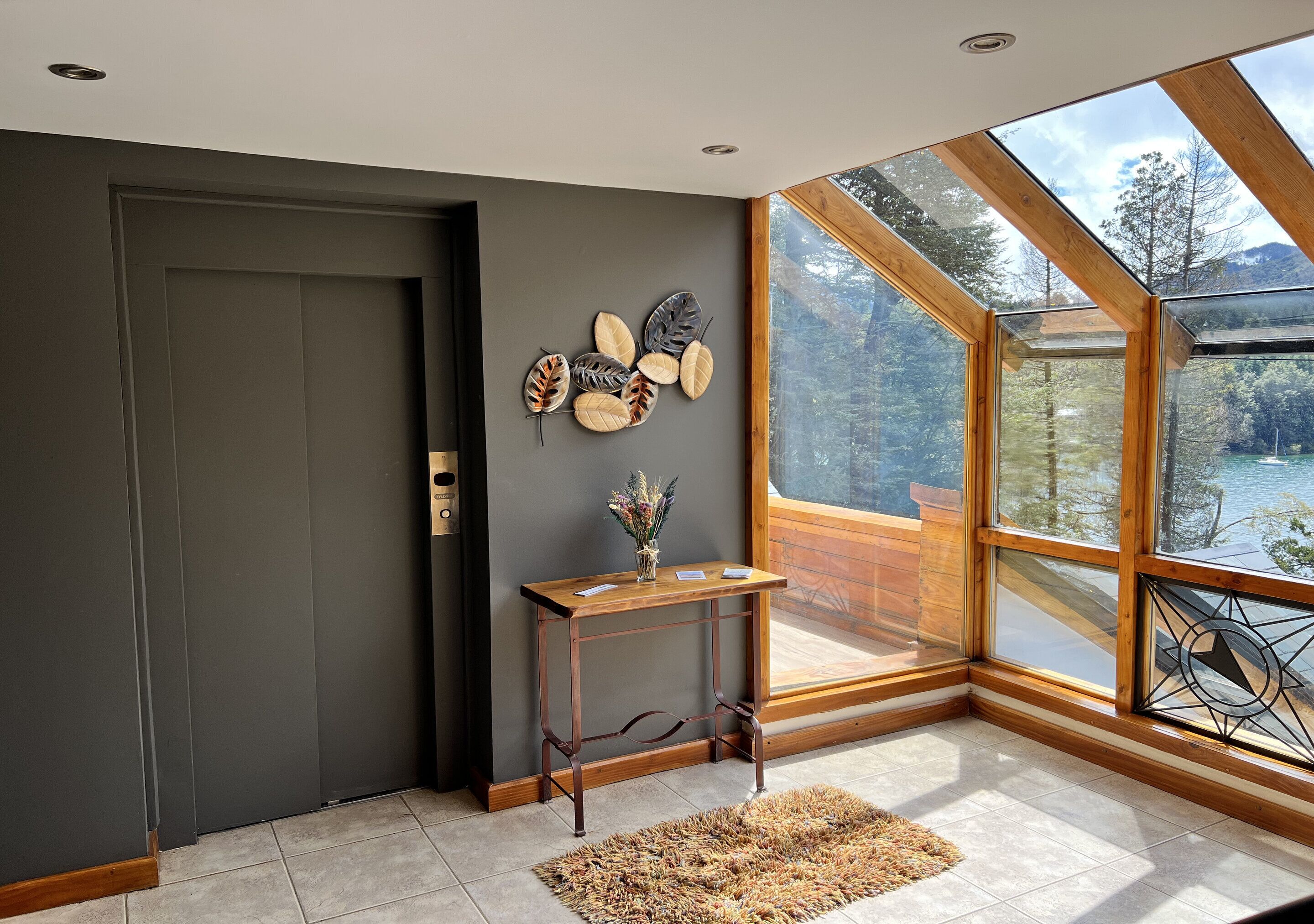 A modern interior with a dark grey door, a wooden console table with decorative leaves, a small vase, and a view of a forest through large windows.