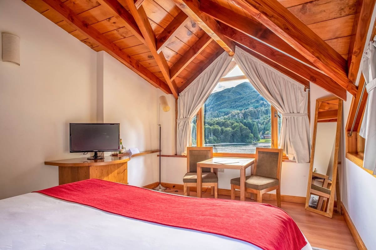 A room with a bed, a desk with a computer, a table with chairs, and a large window with a view of a forested mountain.