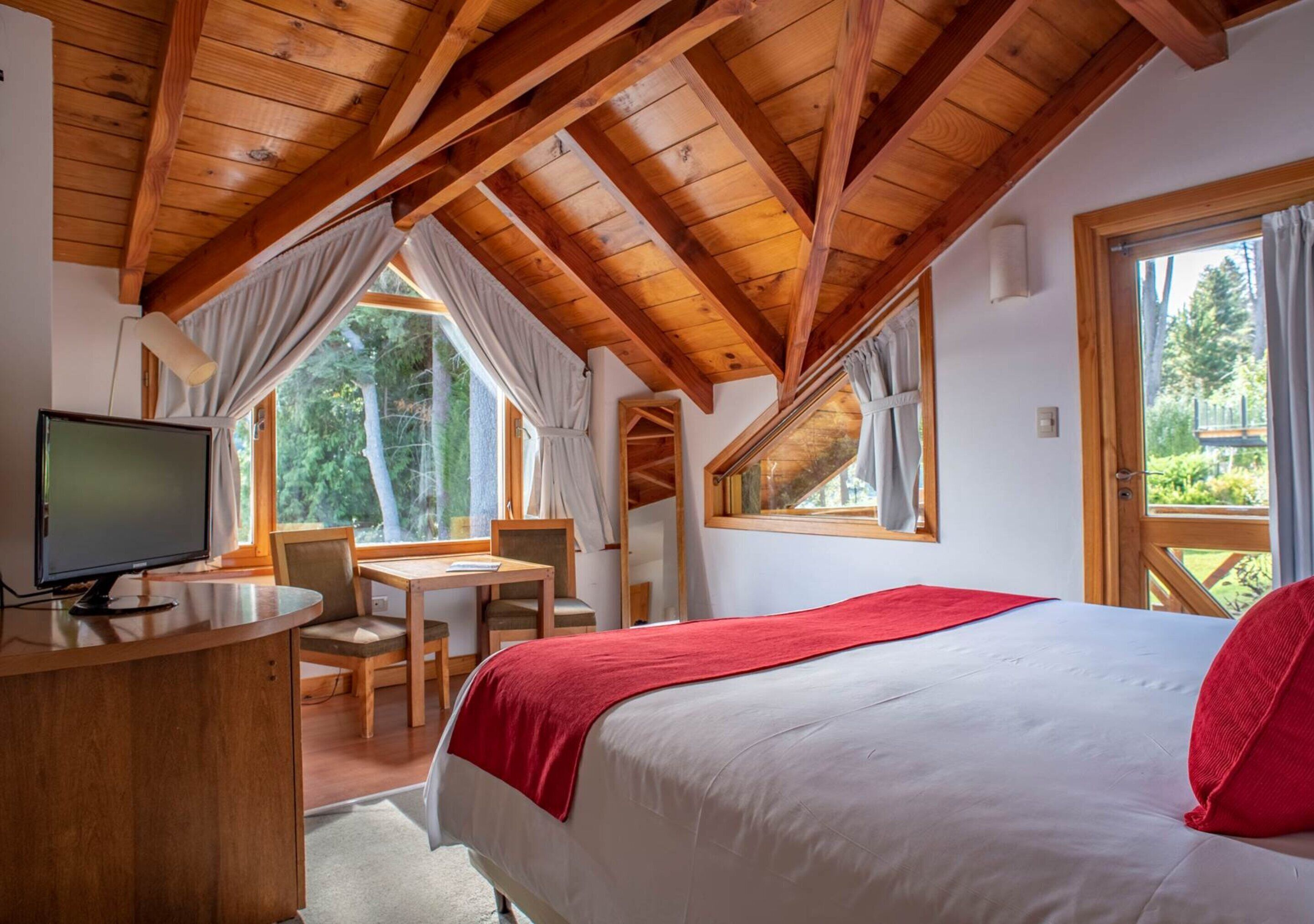 A bedroom with a wooden ceiling, a bed with a red blanket, a desk with a computer, and windows offering a view of trees.