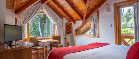 A bedroom with a wooden ceiling, a bed with a red blanket, a desk with a computer, and windows offering a view of trees.
