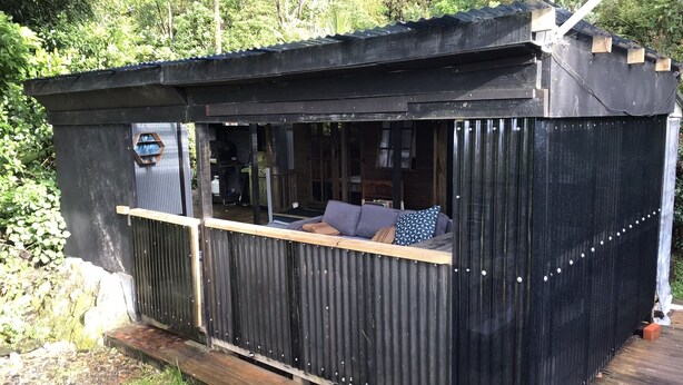 En liten, svart stuga med en träveranda och en sittplats med fönster.
