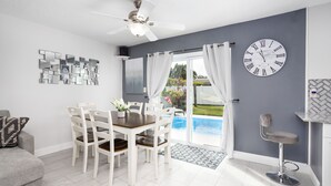 A modern living room with a dining table, chairs, a ceiling fan, a large clock, and a view of a swimming pool. - House in Melbourne With Pool (Melbourne)