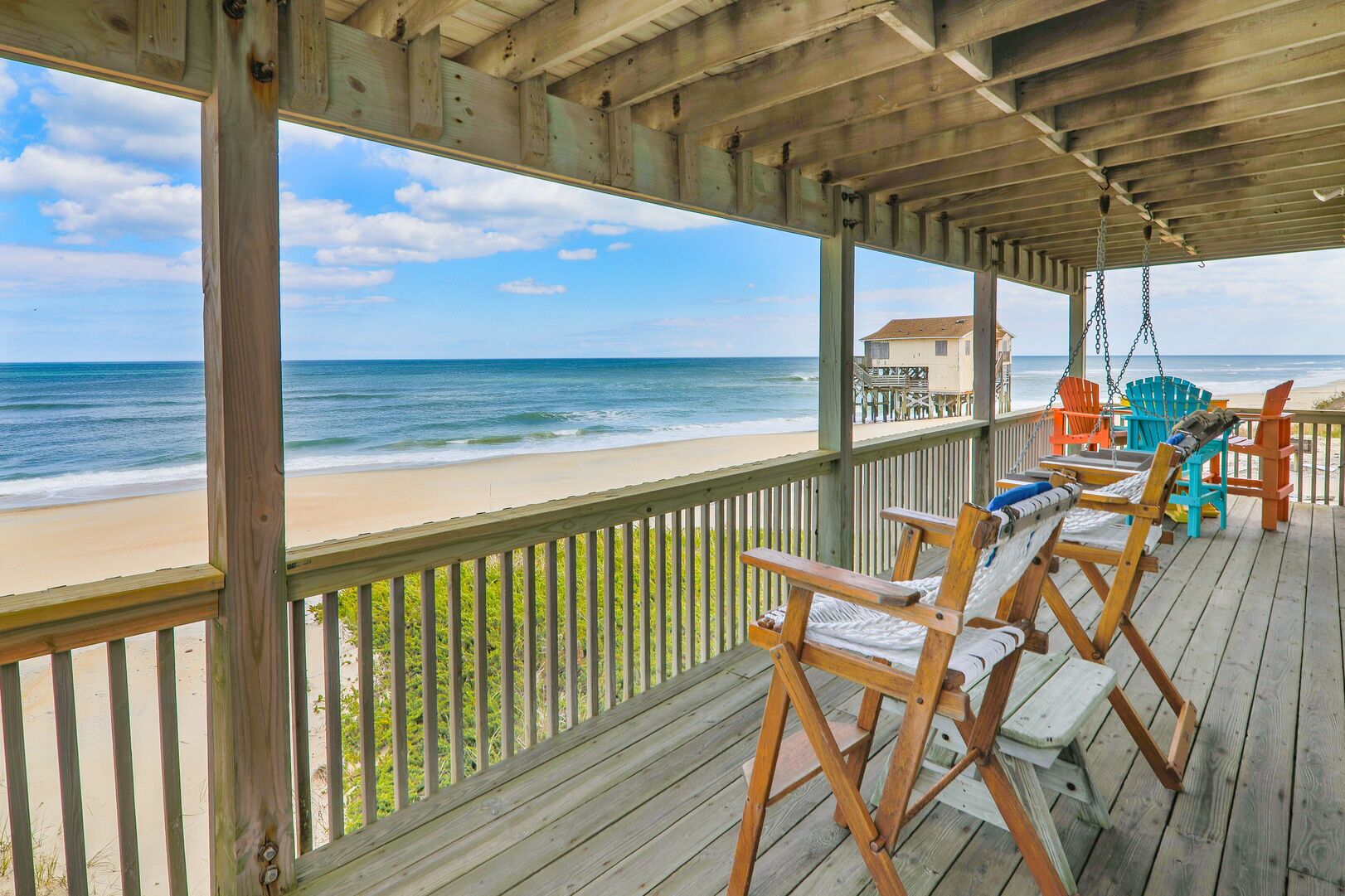 Une terrasse en bois avec des chaises de plage et une balançoire, donnant sur une plage de sable et l’océan.