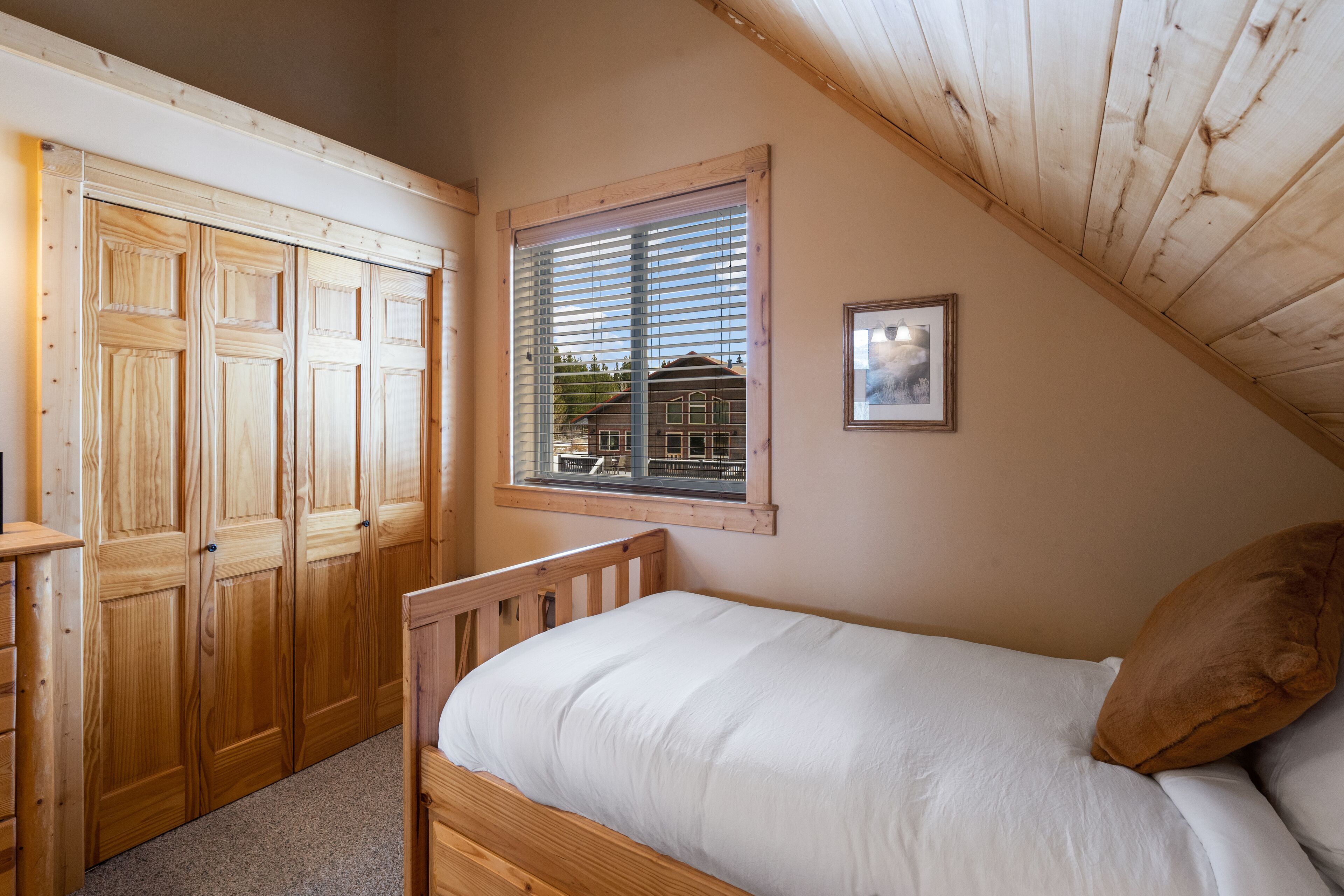 A bedroom with a wooden bed, a window with blinds, and a view of a house outside.