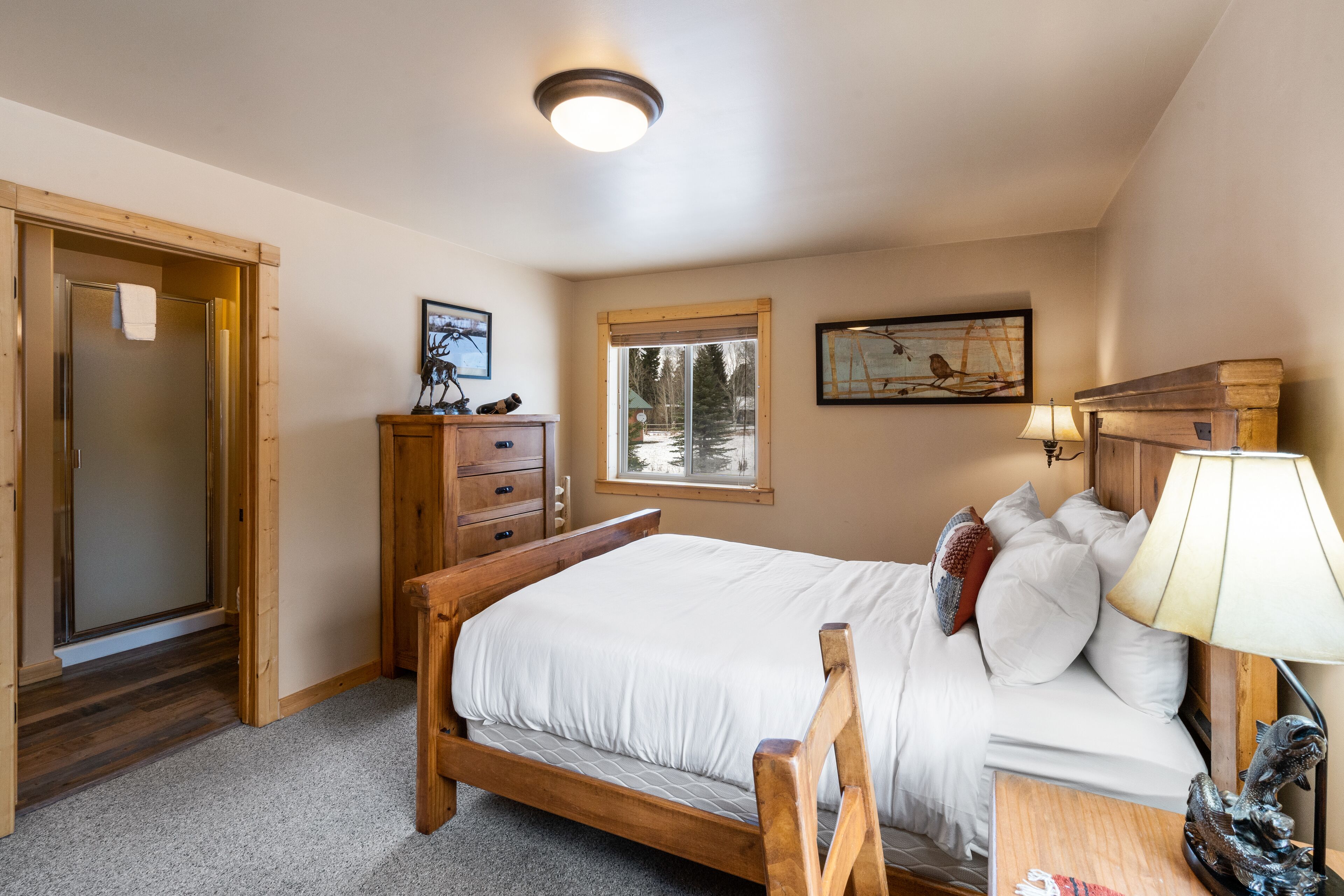 A bedroom with a wooden bed, a nightstand, a lamp, a dresser, and a window with a view of trees.