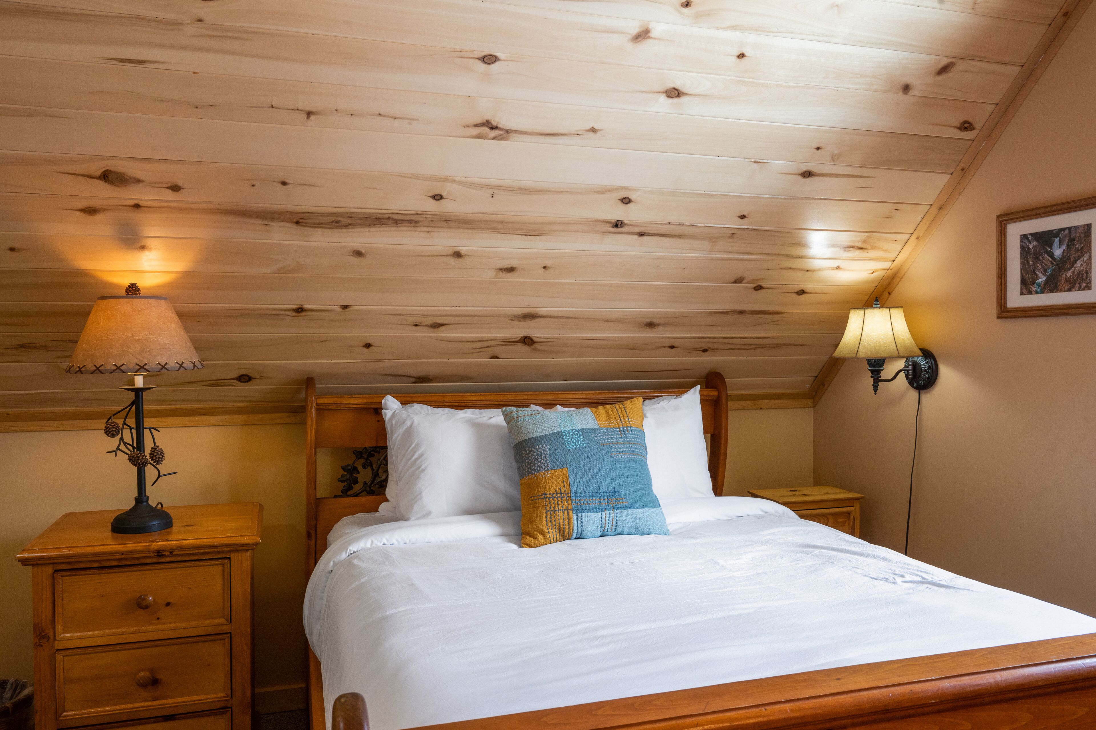 A wooden ceiling, a bed with white and blue pillows, a nightstand with a lamp, and a wall-mounted lamp.