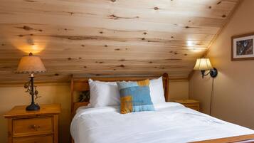 A wooden ceiling, a bed with white and blue pillows, a nightstand with a lamp, and a wall-mounted lamp.