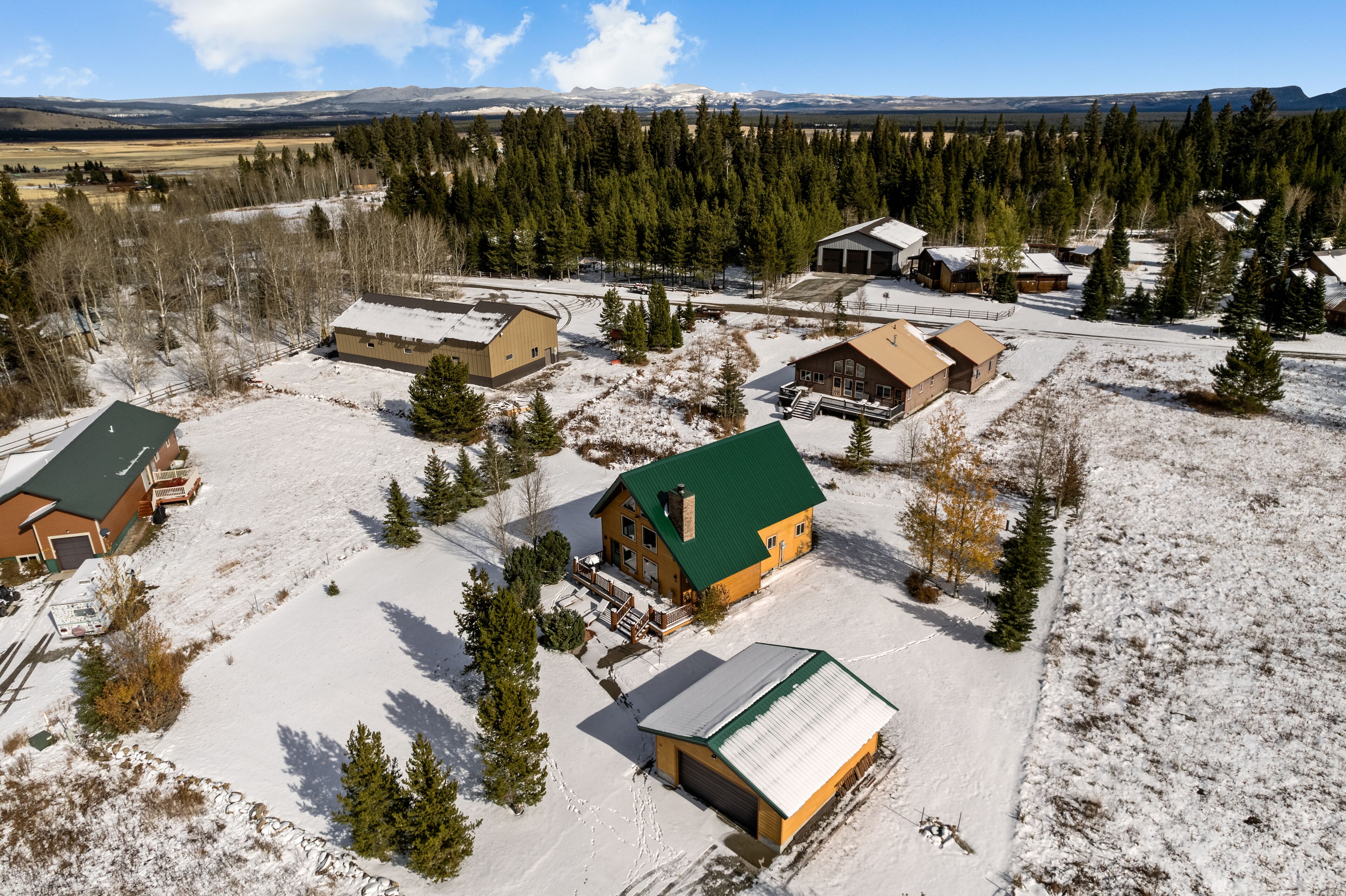 An aerial view of a snow-covered rural area with several buildings, including cabins and a large house with a green roof, surrounded by trees and open fields.