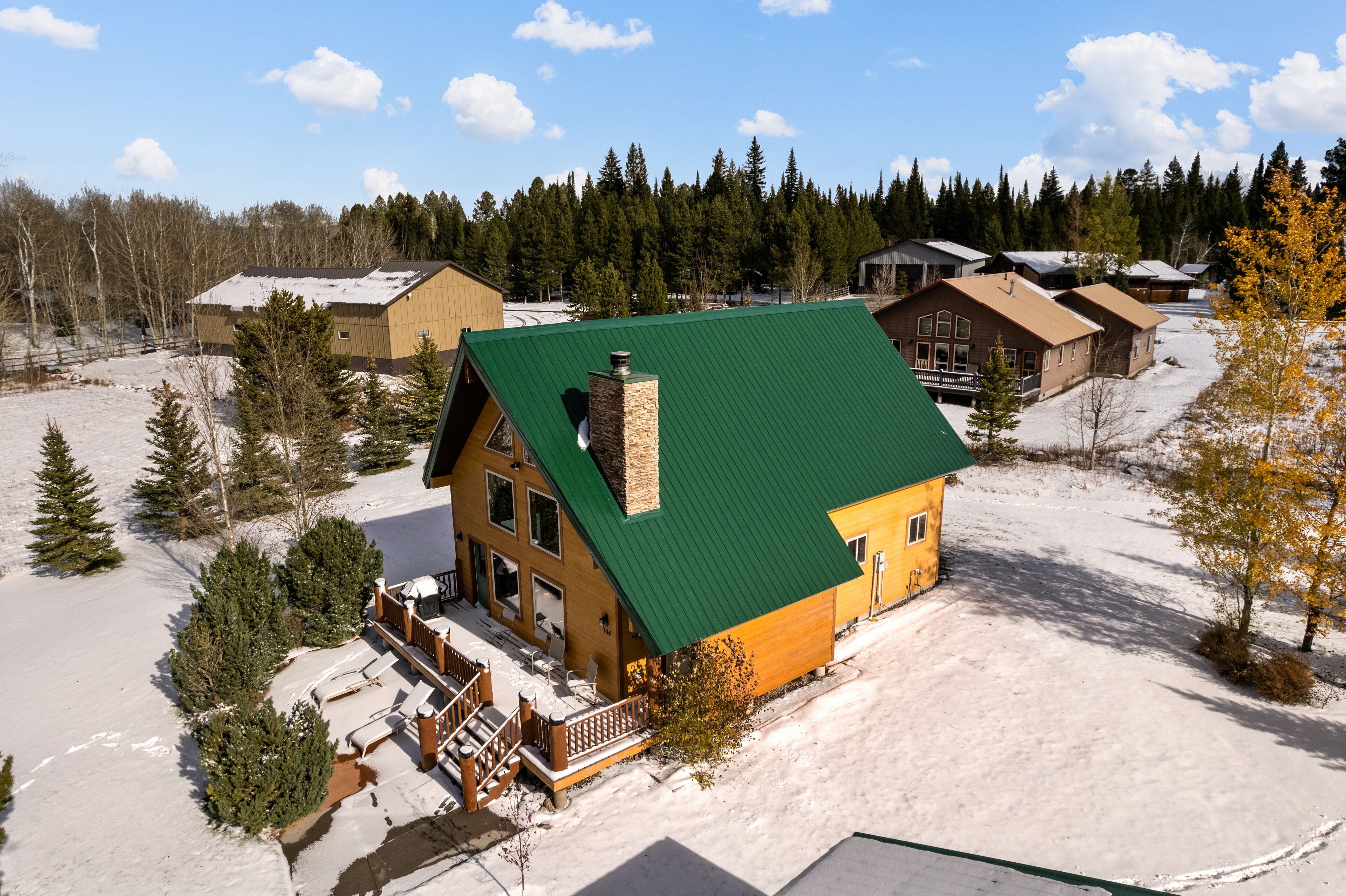 A lodge with a green roof, surrounded by snow-covered ground and trees, featuring a wooden deck and multiple buildings in the background.