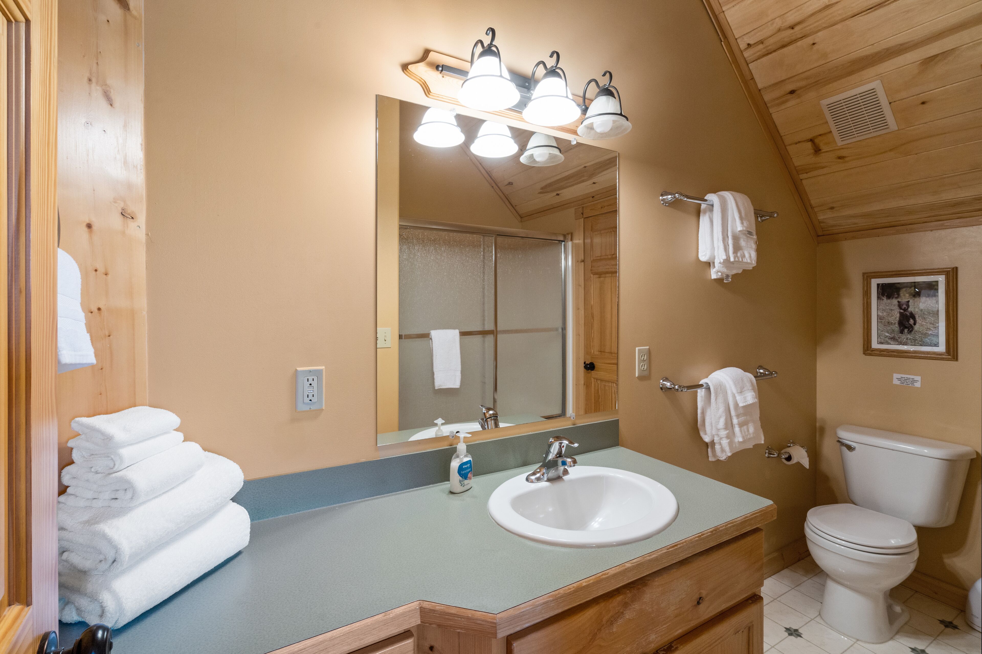 A bathroom with a sink, mirror, and toilet. There are towels stacked on the left, and a picture on the wall.