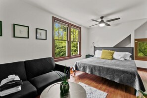 Relax in this serene bedroom with a large bed, ceiling fan, and a sofa, offering a view of lush greenery outside.
