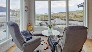 Una habitación con dos sillas grises, una mesita redonda con un jarrón de flores y vistas a un paisaje rocoso a través de amplios ventanales.