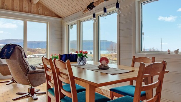 Un comedor con mesa y sillas de madera, ventanas amplias con vistas al exterior y un jarrón con flores sobre la mesa.