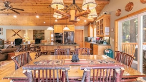 Interior rústico de una cabaña con una mesa de comedor de madera rodeada de sillas, una zona de cocina con electrodomésticos modernos y un gran ventanal con vistas al exterior.