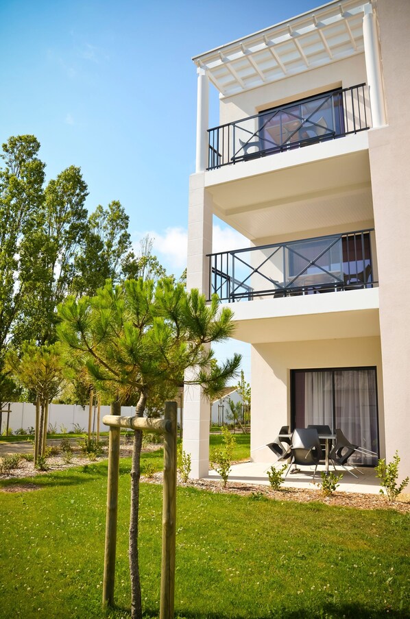 A modern two-story building with a balcony on the upper floor, a patio area with outdoor furniture, and a well-maintained lawn with trees.