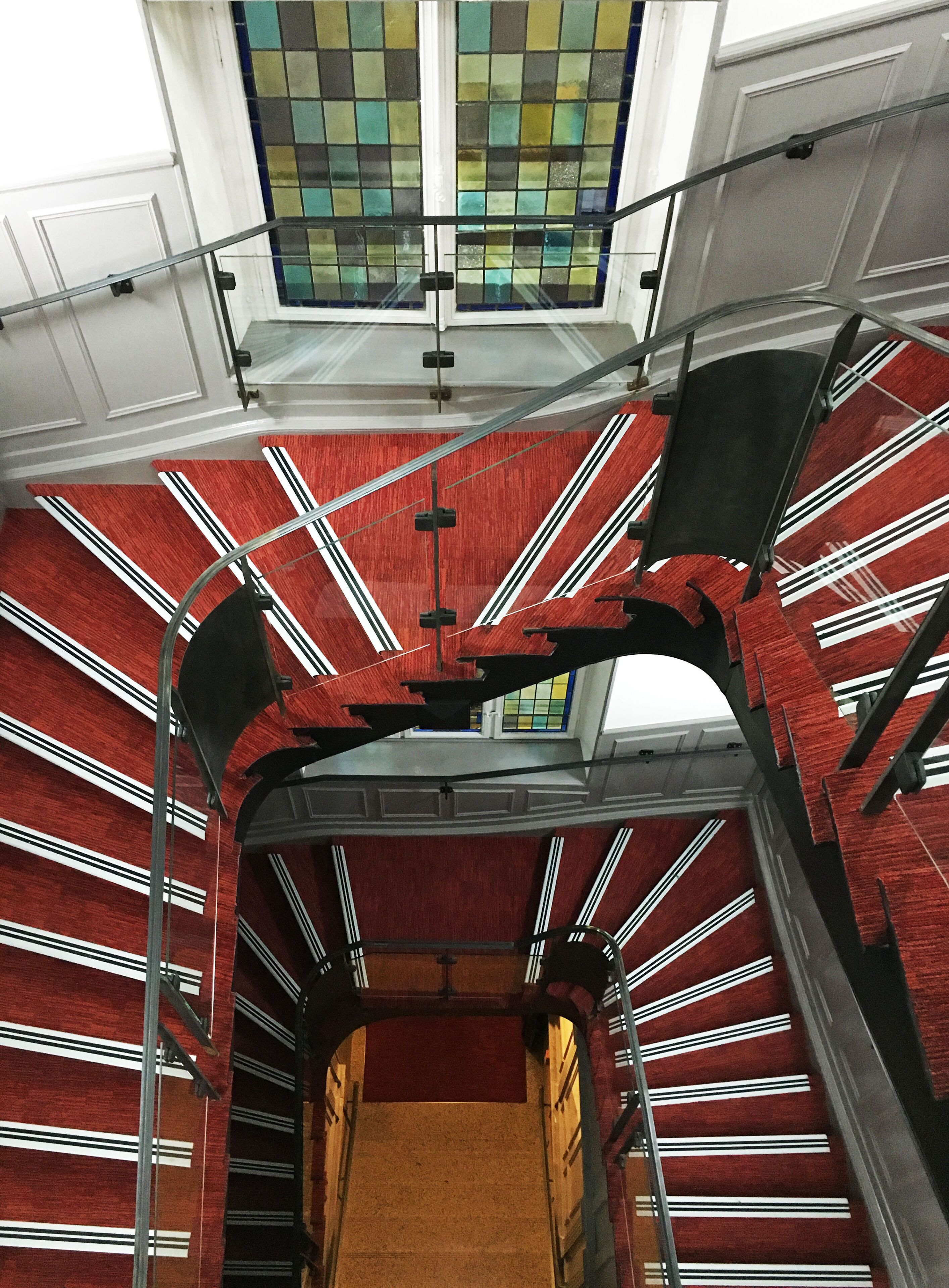 Eine Wendeltreppe mit roten Stufen und weißen Streifen, die zu einem Raum mit Blick auf ein grün-gelbes Buntglasfenster führt.