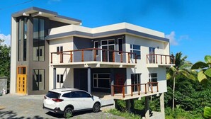Une maison moderne à deux étages, dotée d’un balcon, avec une voiture garée devant et entourée d’une végétation luxuriante.