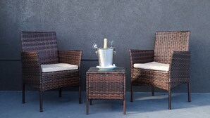 Deux fauteuils en osier avec des coussins, un seau en métal et une petite table, le tout placé contre un mur à texture.