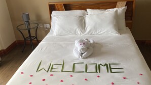 Un lit bien fait, avec une serviette en forme de lapin, une pancarte « BIENVENUE » faite de feuilles vertes et des pétales de fleurs roses éparpillés.