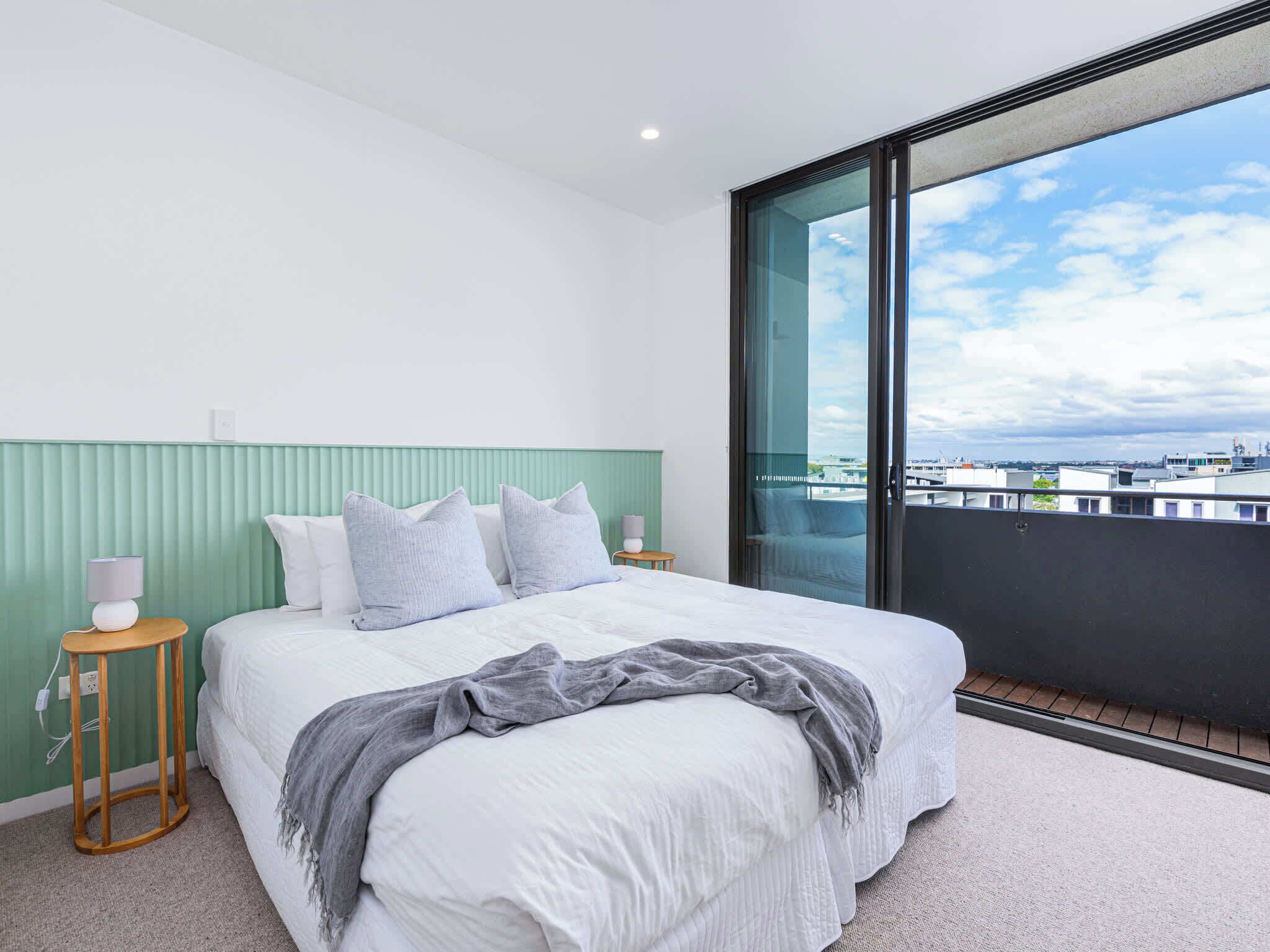 Une chambre moderne avec un grand lit, une tête de lit verte, une table de chevet et une vue sur les immeubles par une grande fenêtre.