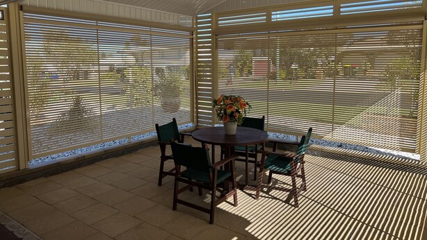 Un patio techado con una mesa redonda y sillas, ventanas grandes con persianas y una planta en maceta.